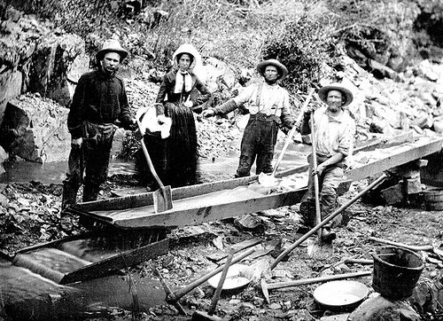 Historic 1850 photograph of a woman and three men panning for gold beside a flume during the California gold rush
